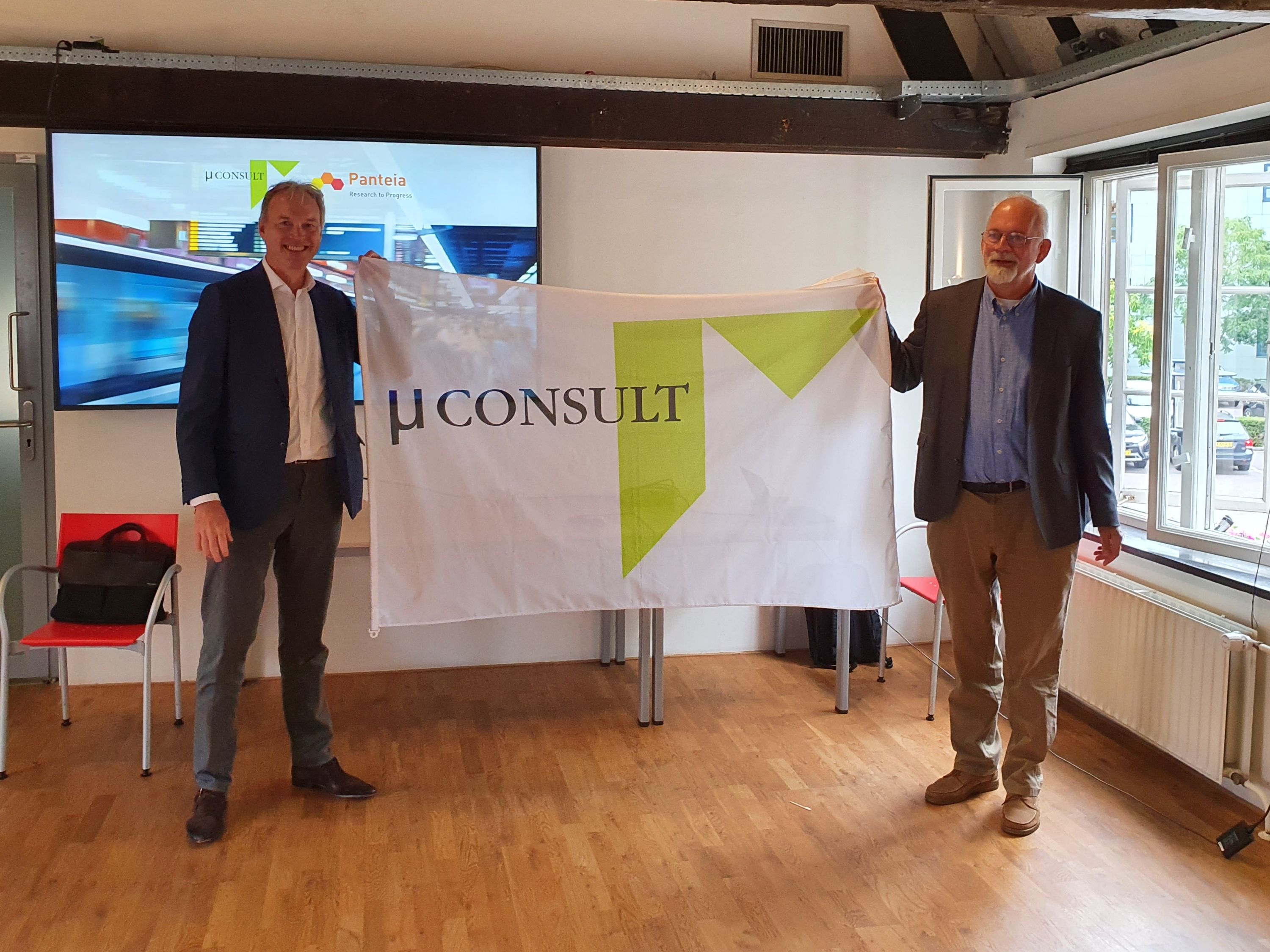 foto van de twee directeuren die een vlag ophouden van het logo van MuConsult
